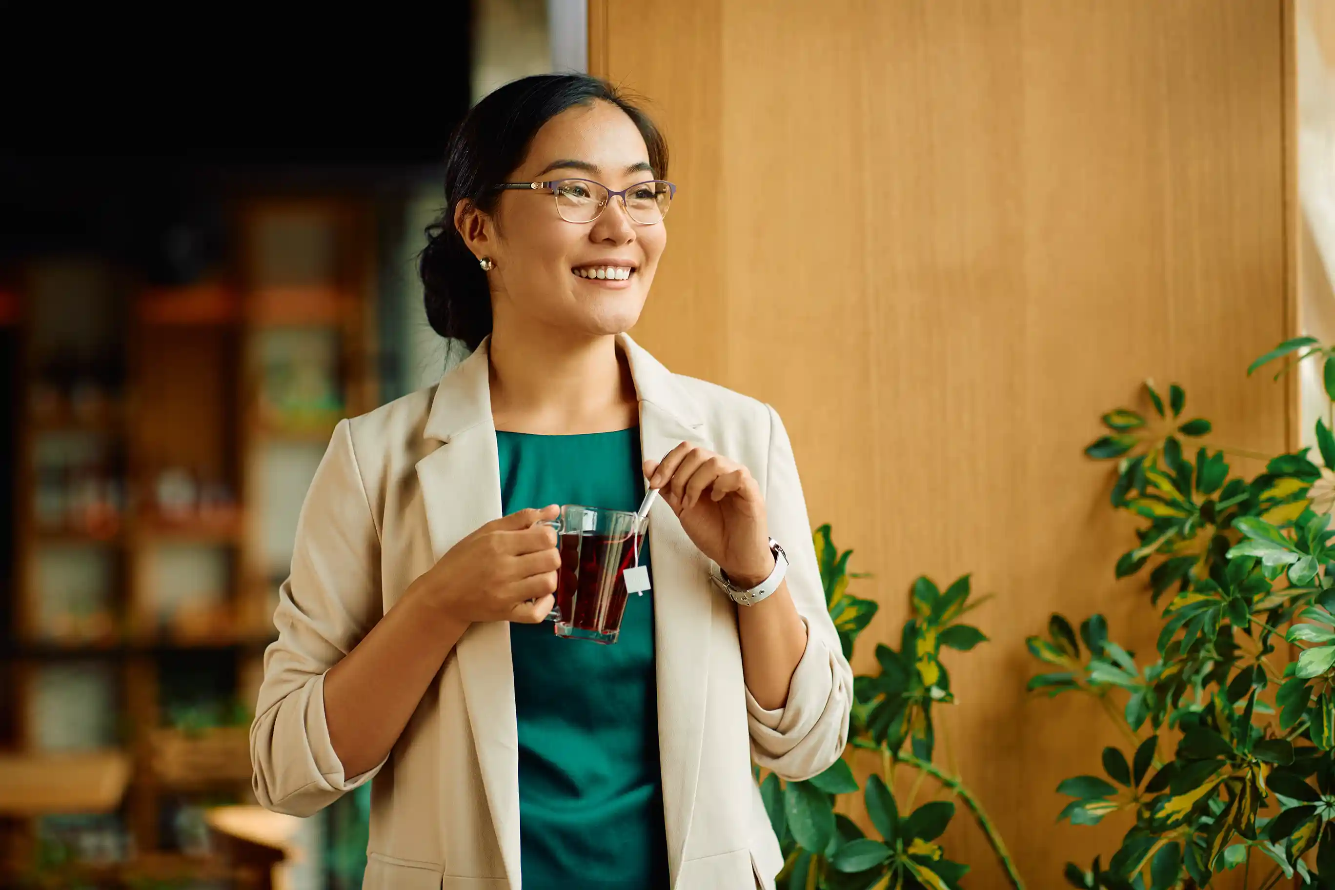Mulher sorridente com óculos e blazer bege, segurando uma xícara de chá em um ambiente de escritório moderno com plantas ao fundo.