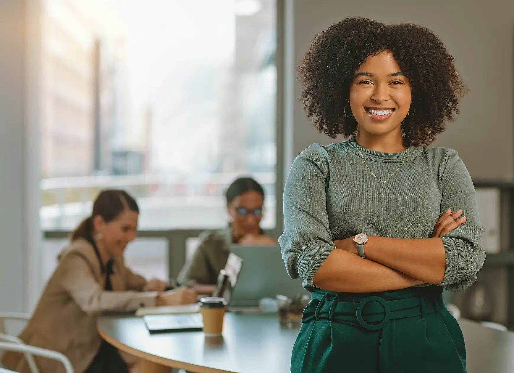Retrato de uma mulher negra sorridente com cabelos cacheados e braços cruzados; ao fundo, colegas trabalham em uma mesa de reunião.