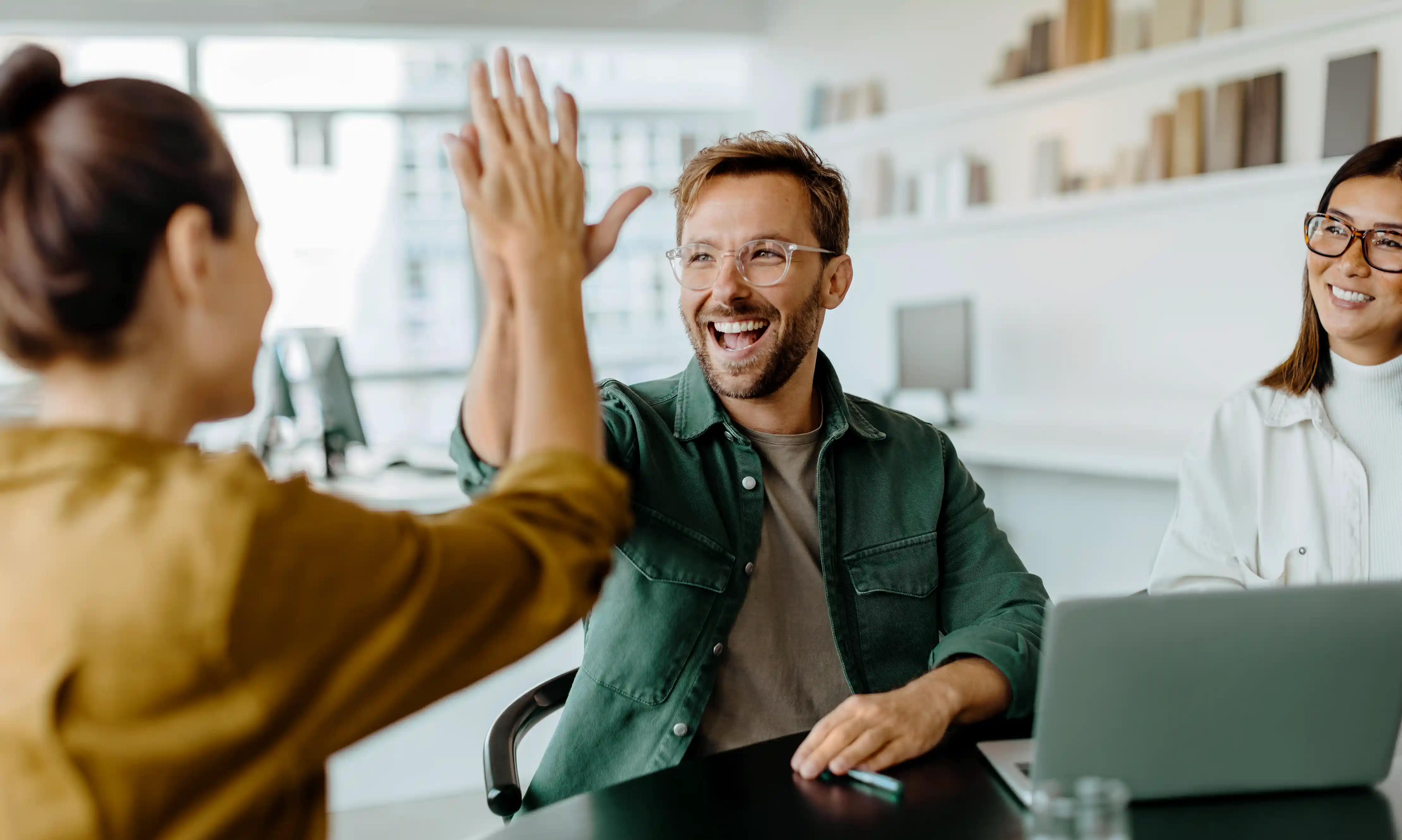 Um homem com óculos e camisa verde comemorando com um "high five" (toca aqui) com uma colega durante uma reunião de equipe.